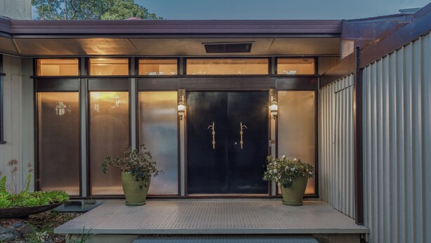 A modern building entrance with a dark double door, two potted plants, and a small outdoor seating area.