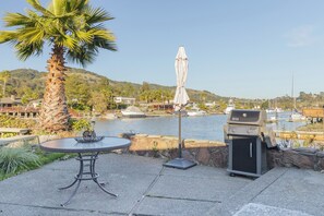 Eine Terrasse mit Tisch und Grill, mit Blick auf einen Yachthafen, wo Boote liegen, und einen Hügel im Hintergrund.