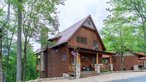 Une cabane rustique en rondins de bois, avec des fondations en pierre, une véranda dotée de sièges d’extérieur et un drapeau américain.