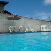 Piscina com cadeiras de descanso brancas e uma mesa, e uma parede revestida com telhado vermelho.