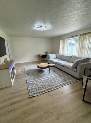 Una sala de estar con un sofá, dos mesas redondas de madera, un tapete estampado, un televisor y una pequeña mesita auxiliar.