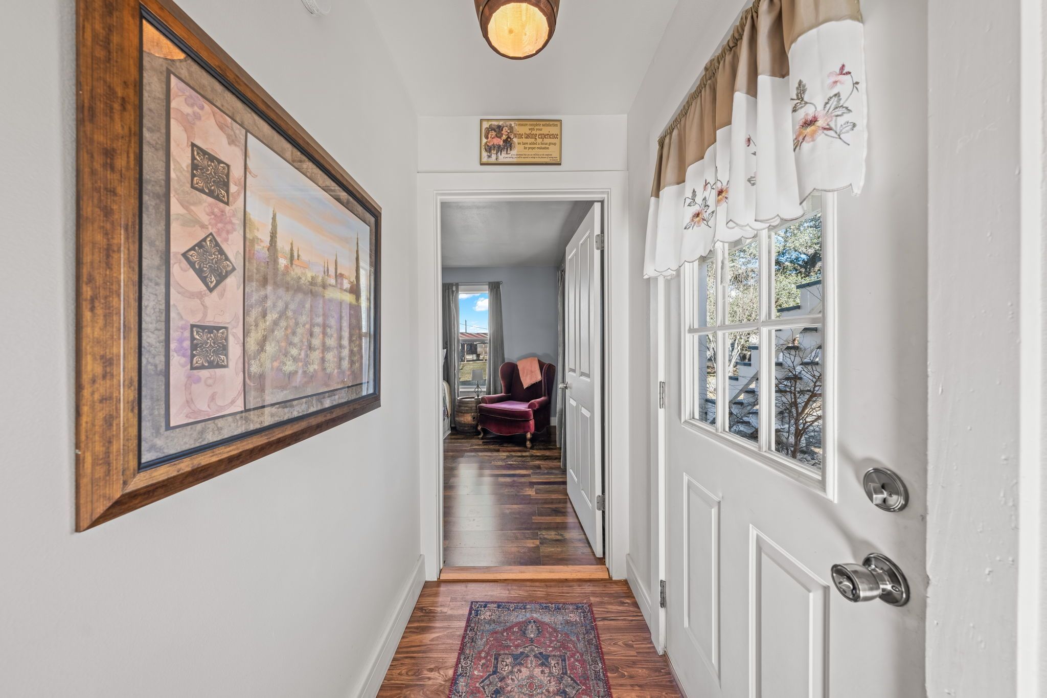 A hallway with a framed artwork on the left, a red chair in the background, and a door with decorative curtains on the right.