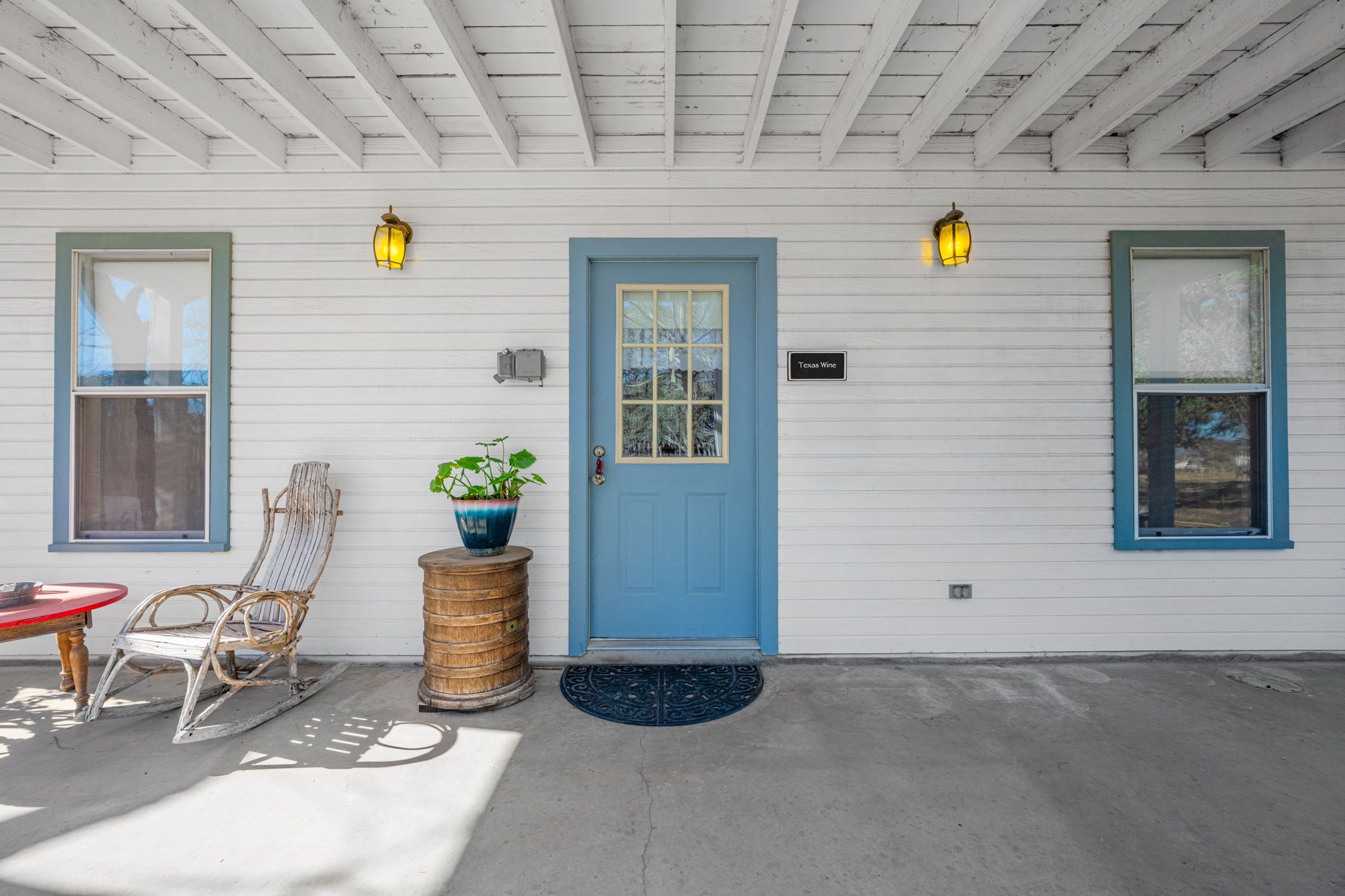 A house with a blue door, two windows, a porch with a chair and a table, and a potted plant.