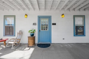 A house with a blue door, two windows, a porch with a chair and a table, and a potted plant.