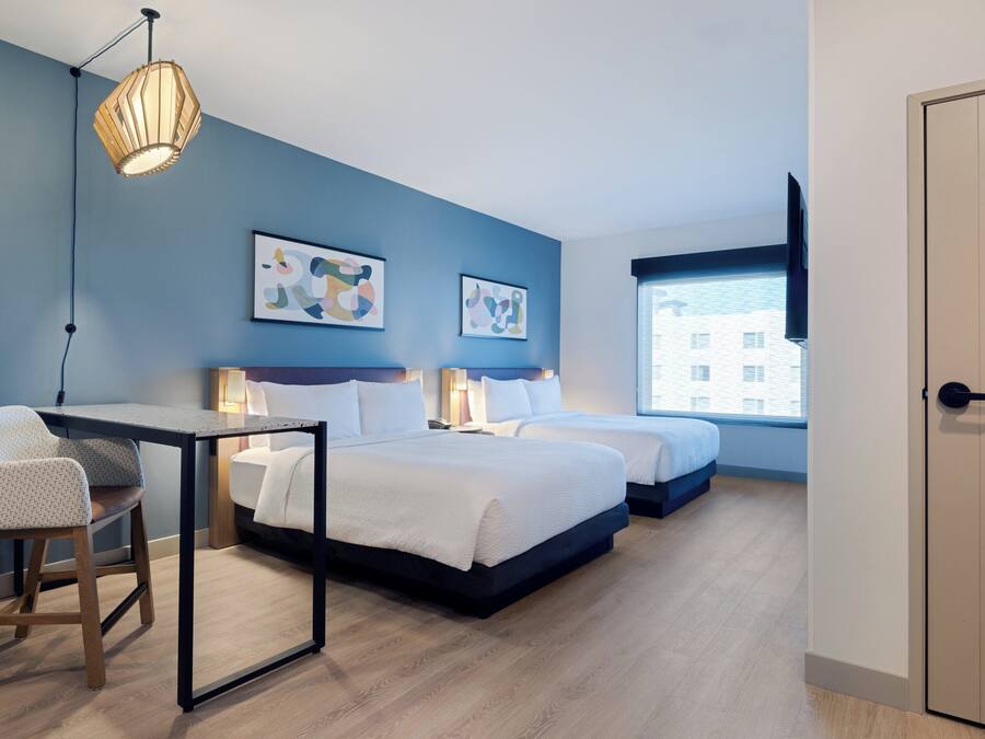 Habitación de hotel moderna con dos camas, un escritorio y una silla. Hay pinturas abstractas en la pared y una ventana con vistas a edificios.