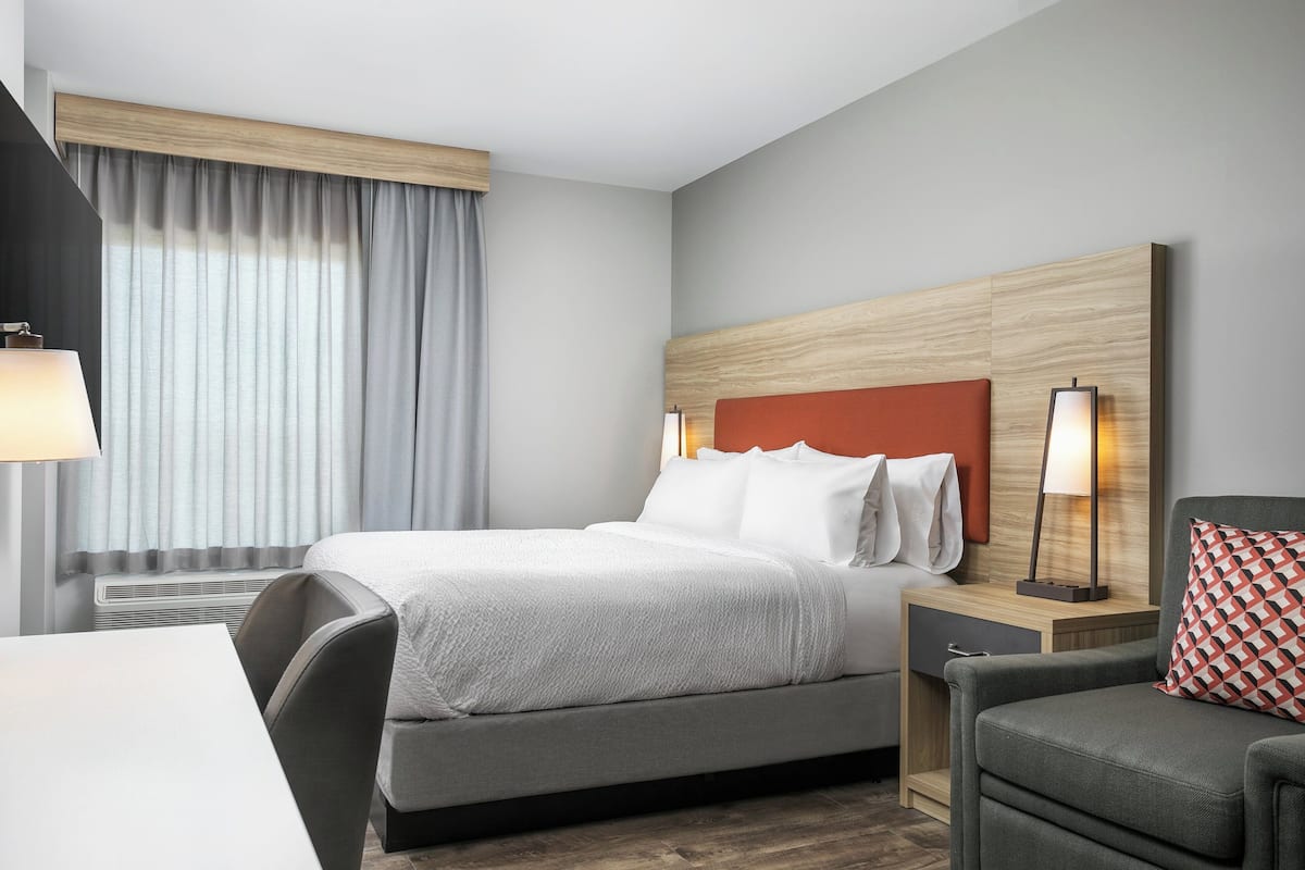 Moderni hotellihuone, jossa on sänky, työpöytä, tuoli ja yöpöytä, jossa on lamppu.