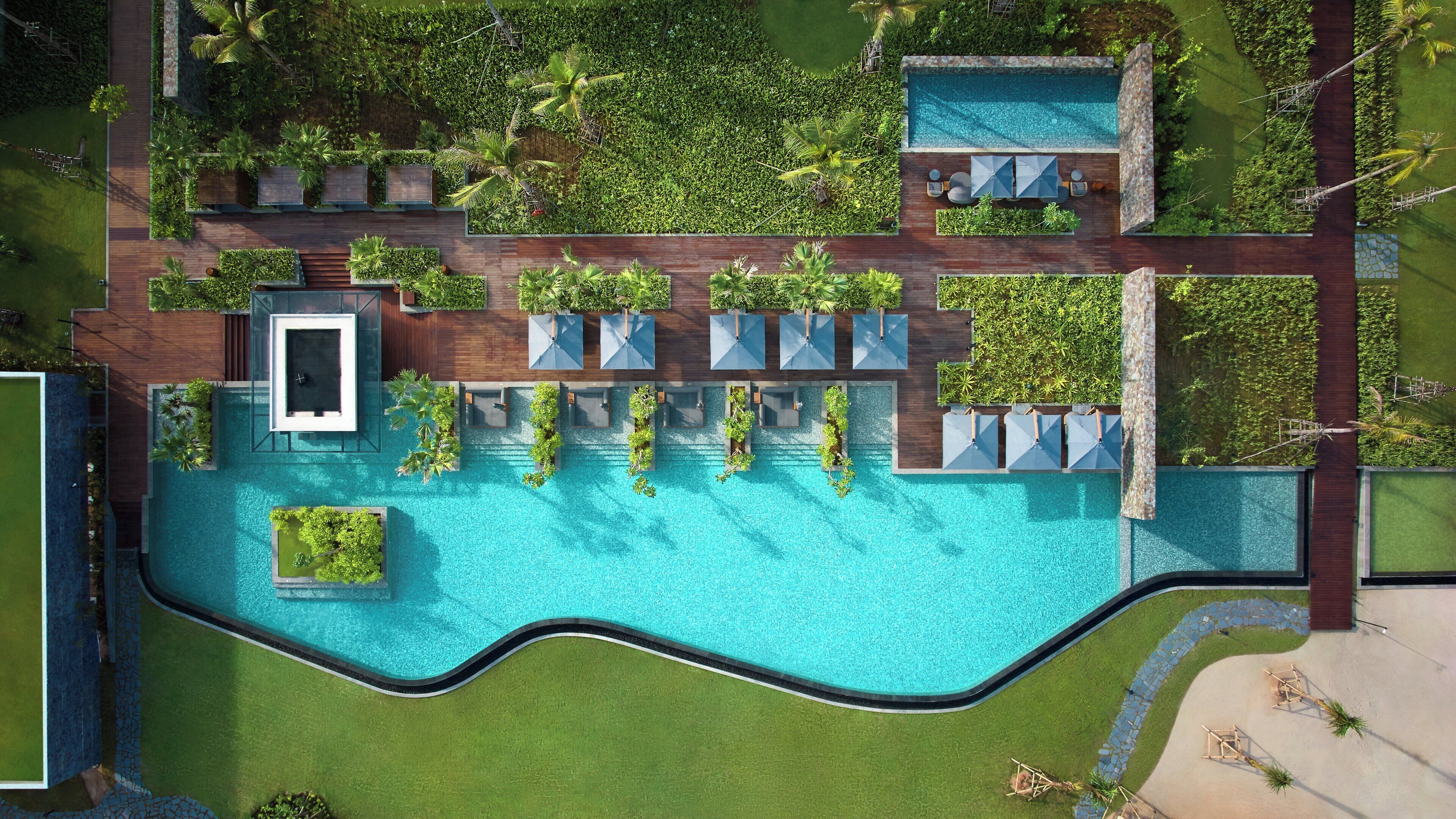 An aerial view of a resort with a large swimming pool, multiple lounge areas with umbrellas, and lush greenery surrounding the facilities.