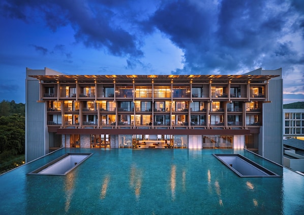 A modern hotel with multiple balconies, a rooftop pool, and a well-lit facade.