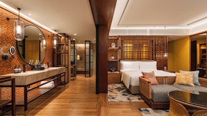 A modern hotel room with a wooden floor, a large bed with a white headboard, a sitting area with a wicker sofa and a glass coffee table, and a bathroom with a large mirror and a marble sink. - Hotel Indigo Bintan Lagoi Beach by IHG (Bintan)