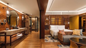 Ein modernes Hotelzimmer mit Holzboden, einem großen Bett mit weißem Kopfteil, einer Sitzecke mit einem Korbssofa und einem Glastisch sowie einem Badezimmer mit großem Spiegel und Marmorwaschbecken.