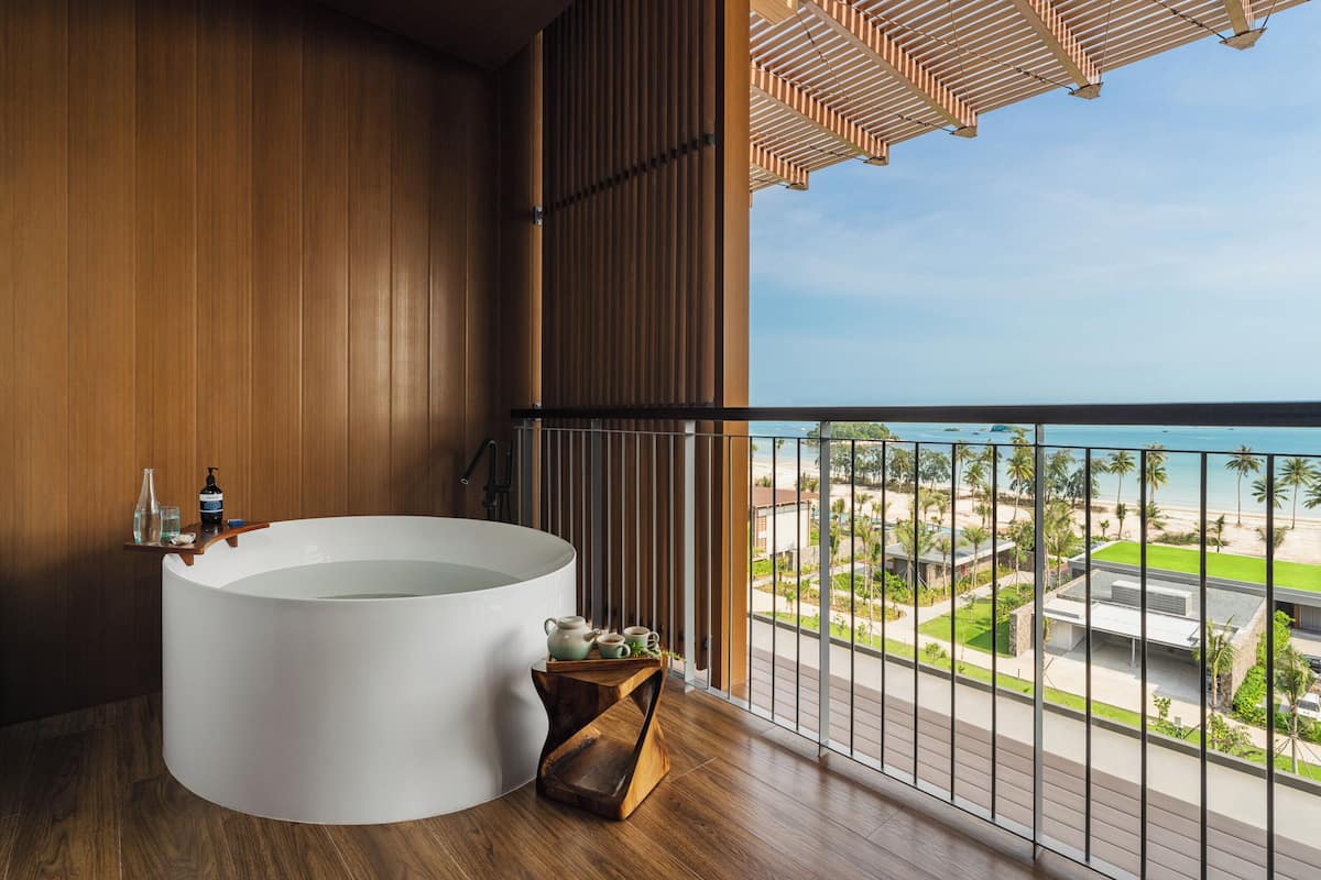 Ein modernes Badezimmer mit einer großen Badewanne, Holzboden und Blick auf den Strand und die Resortgebäude.