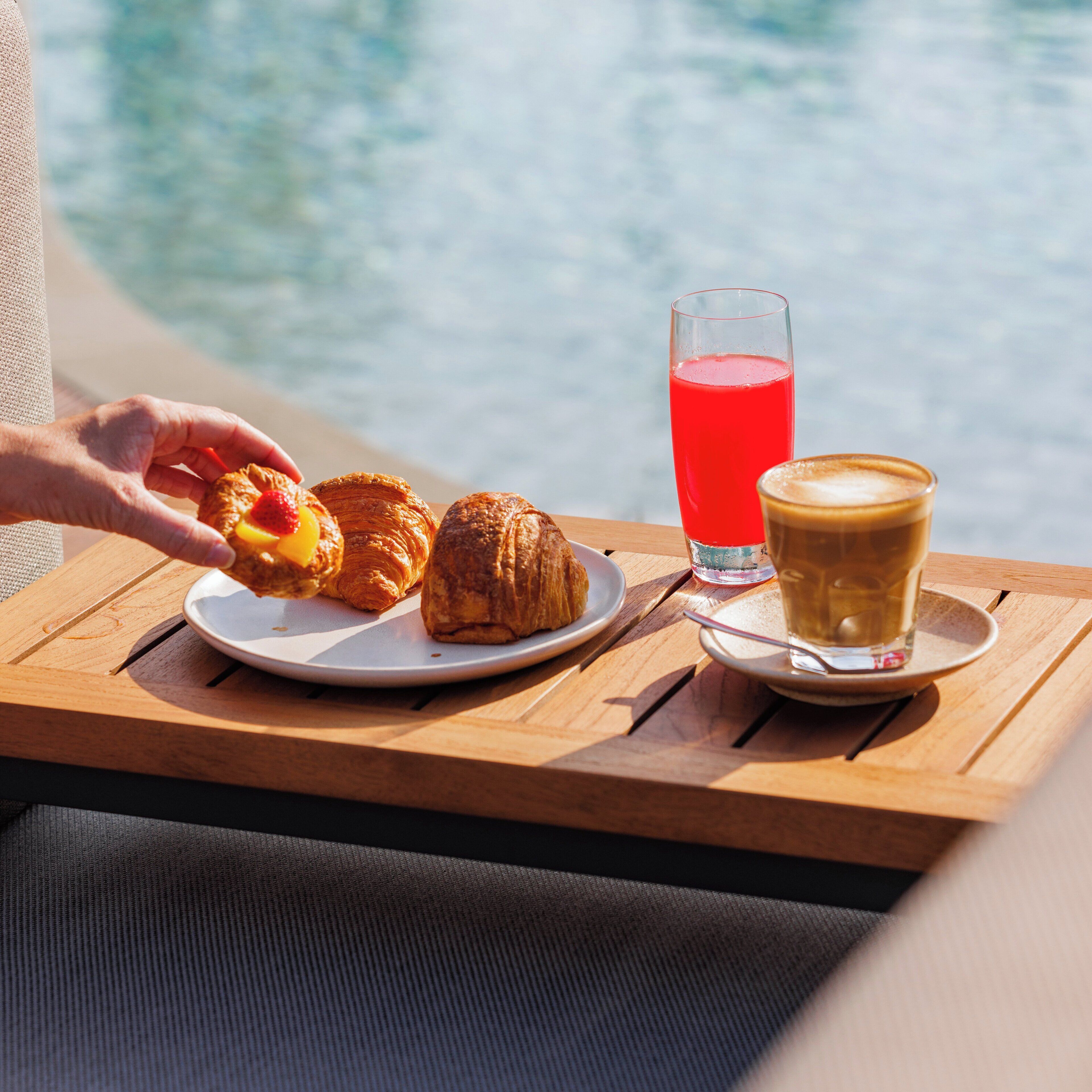 A wooden table with croissants, a glass of red juice, and a cup of coffee, set against a backdrop of a body of water.