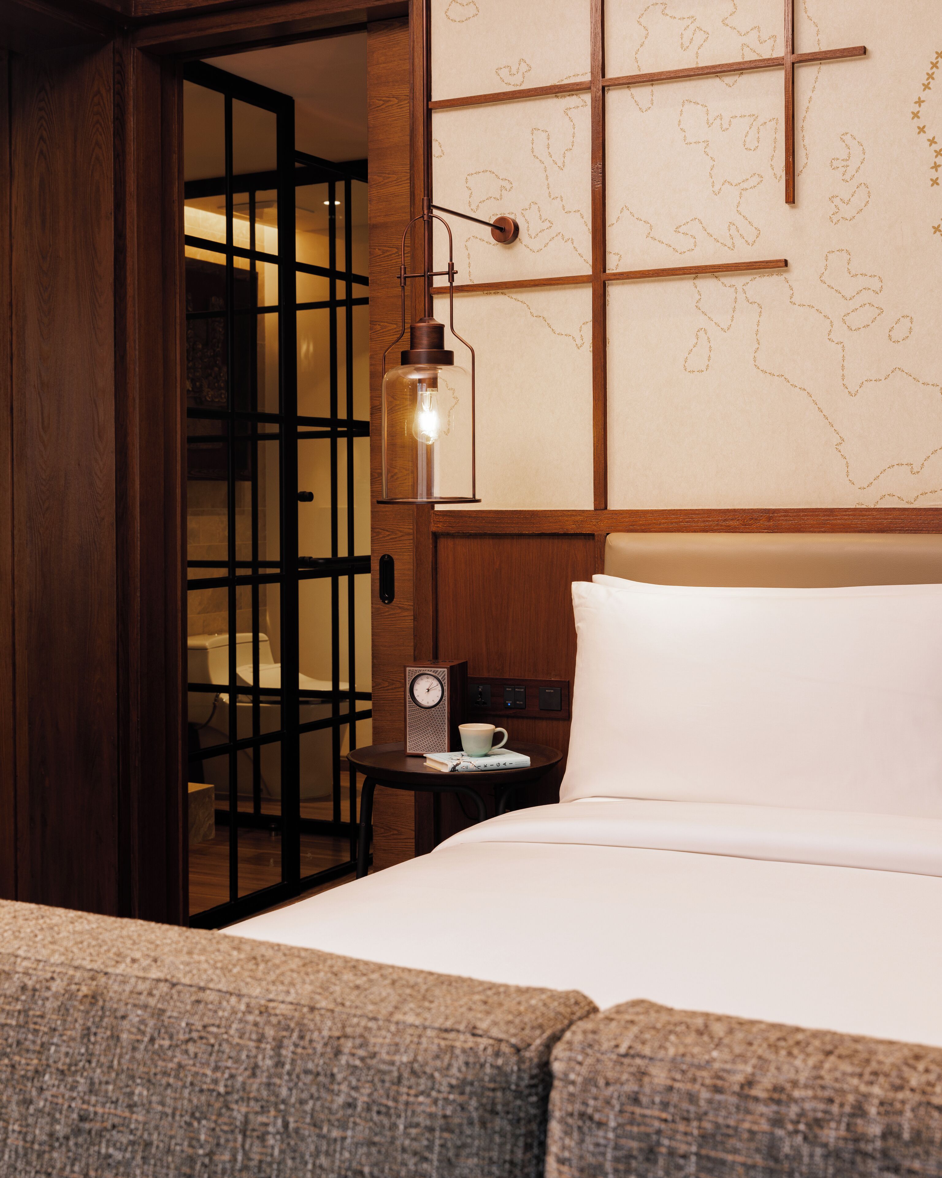 A hotel room with a bed, a small table with a clock and a cup, a wooden door, and a wall with a map design.