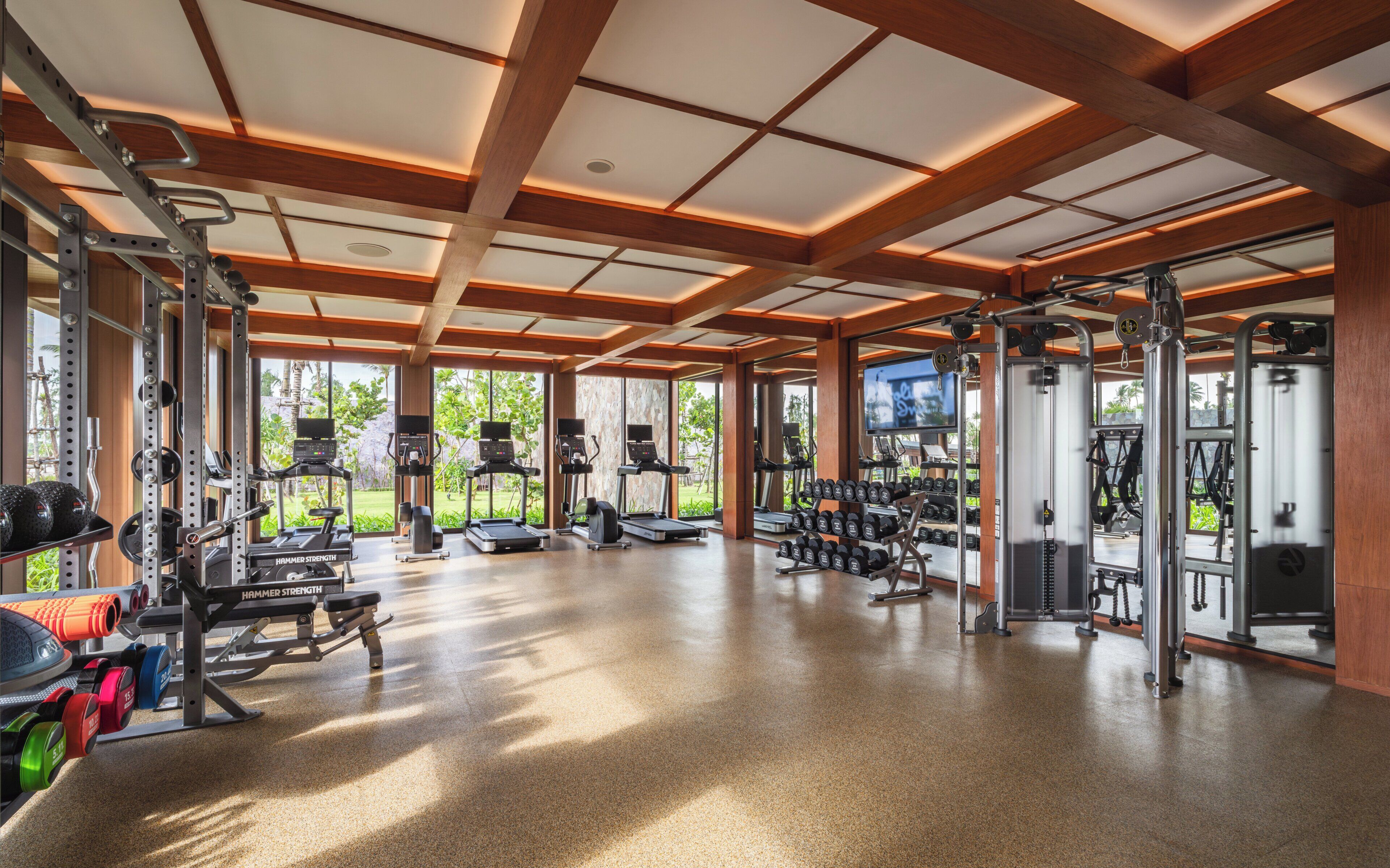 A spacious gym with various exercise equipment, large windows, and a polished floor.