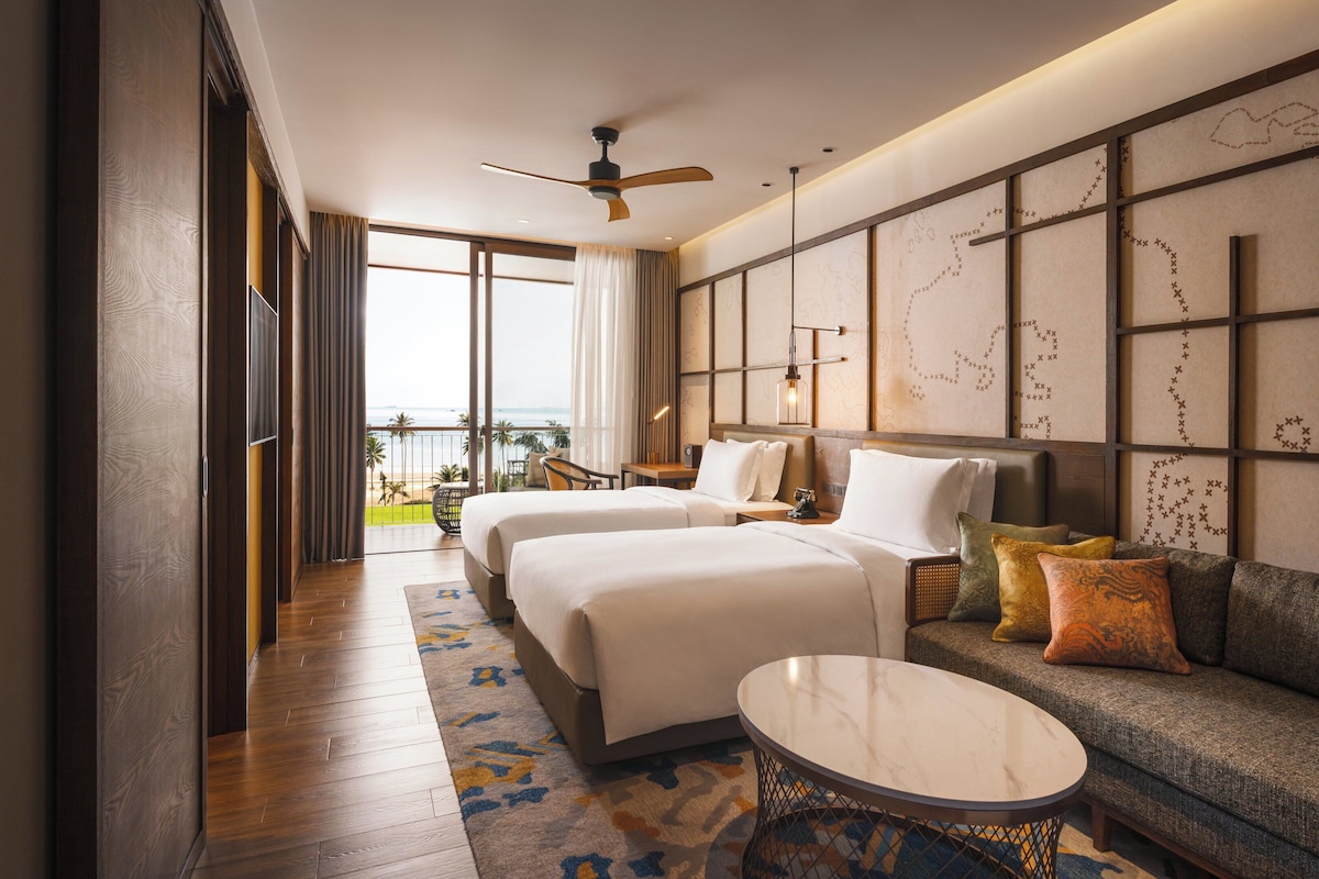 A hotel room with two beds, a sofa, a small table, and a view of the beach.