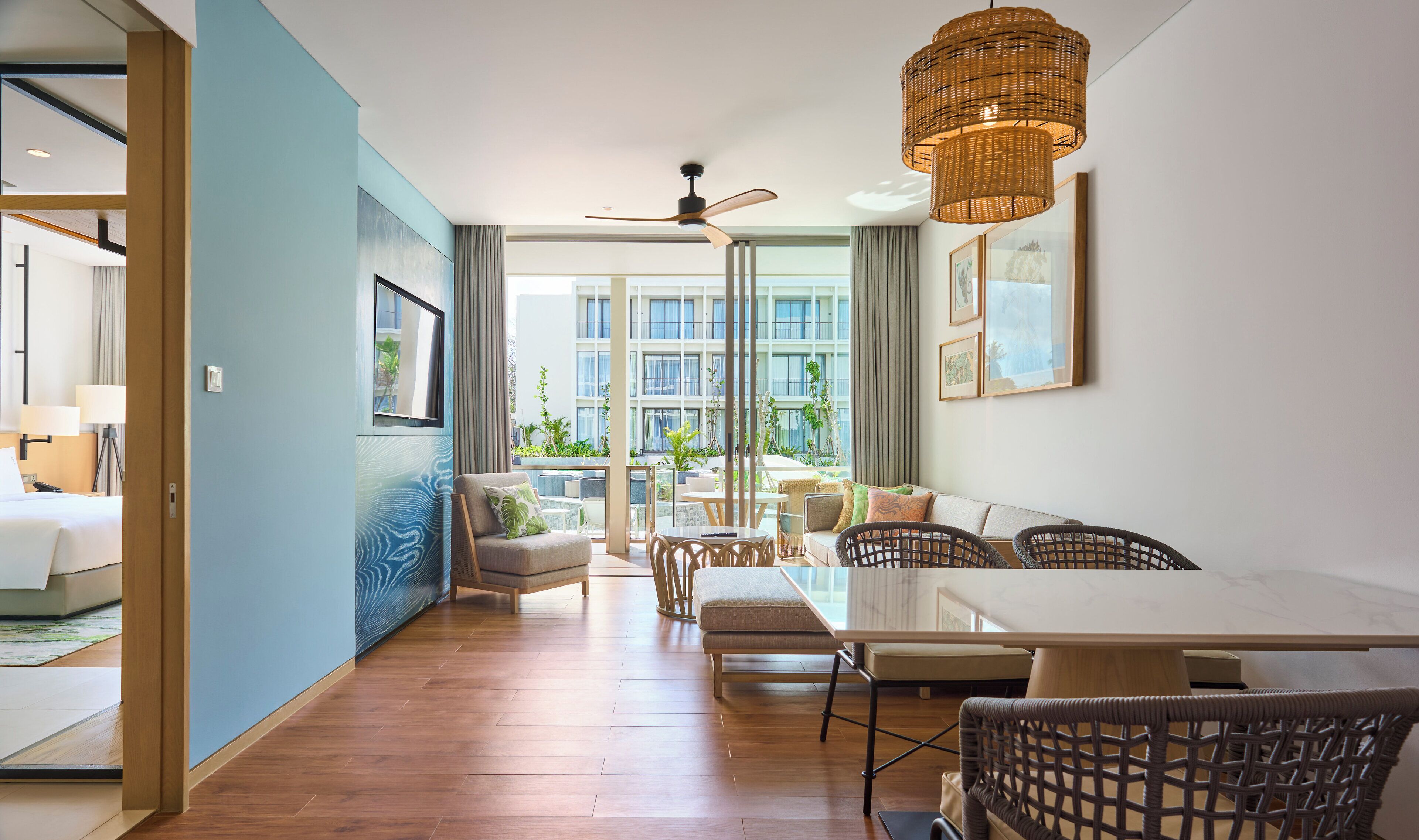 Quarto de hotel moderno com uma janela grande, área de refeições com cadeiras de vime, ventoinha de teto e vista para o exterior do edifício.