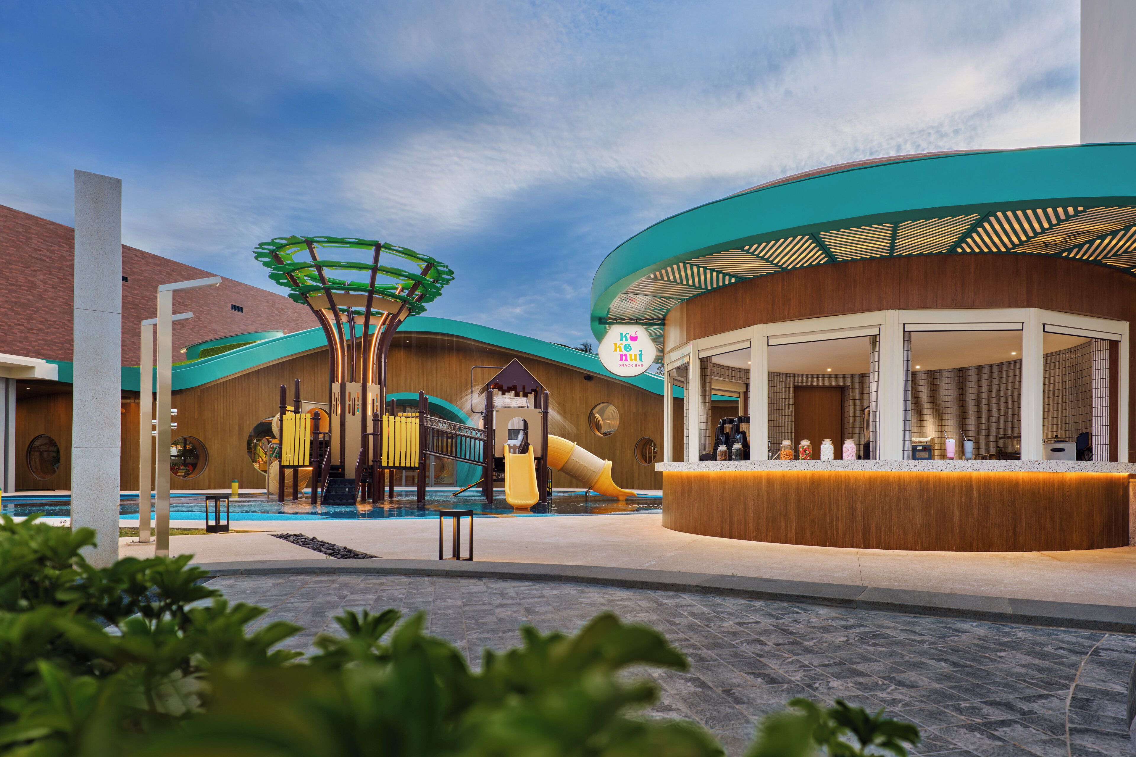 A modern hotel lobby with a pool area, a children's play structure, and a reception desk.