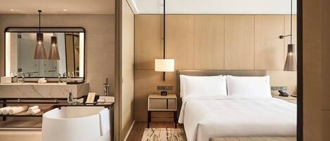 A modern hotel room with a large bed, a round bathtub, a vanity with a mirror, and a bedside table with a lamp.