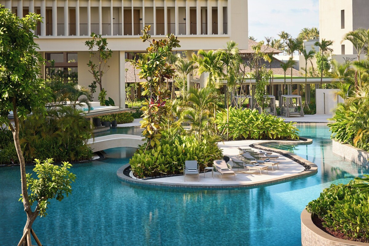 A pool area with lounge chairs and a bridge, surrounded by lush greenery and palm trees.