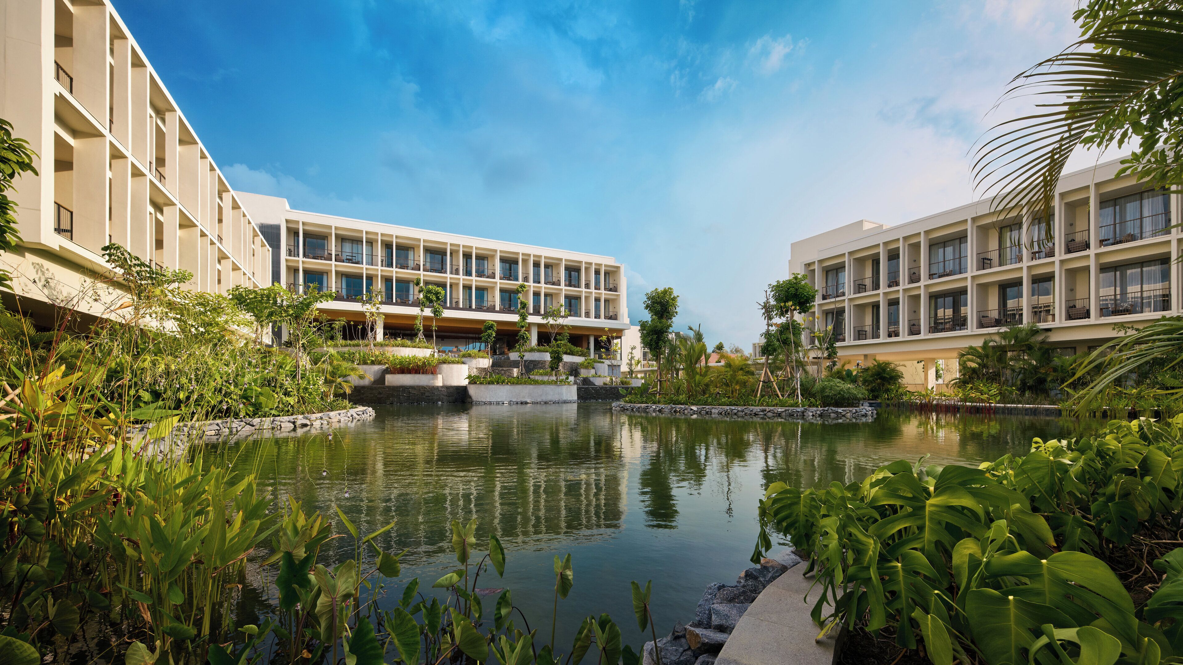 Un hôtel moderne doté d’une cour intérieure où se trouve un bassin d’eau aux reflets changeants, entouré d’une végétation luxuriante et de bâtiments à plusieurs étages dotés de balcons.