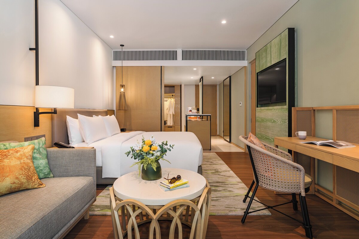 A hotel room with a bed, a sofa, a dining table with chairs, a TV, and a mirror.