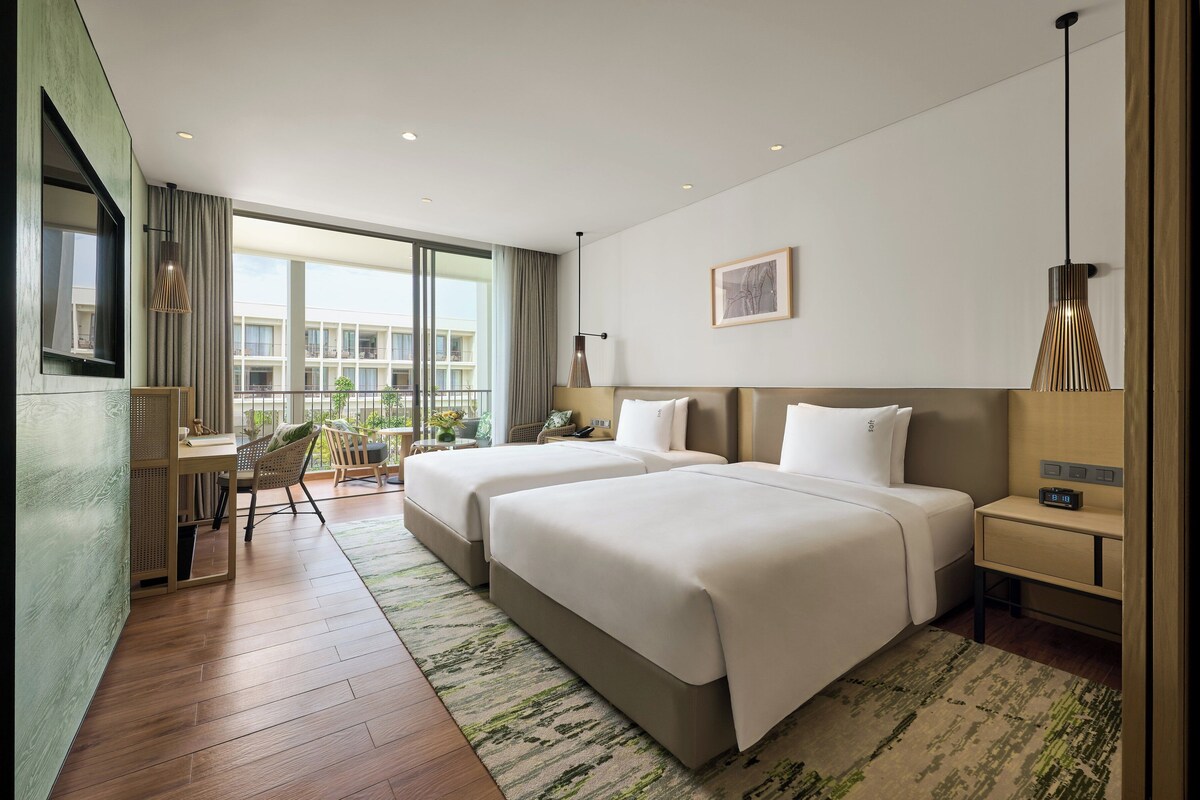 A hotel room with two beds, a desk, a chair, a large window with a view of buildings, and a small table with a lamp.