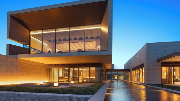Un edifici modern amb façana de vidre, una zona exterior àmplia amb seients i una entrada ben il·luminada.