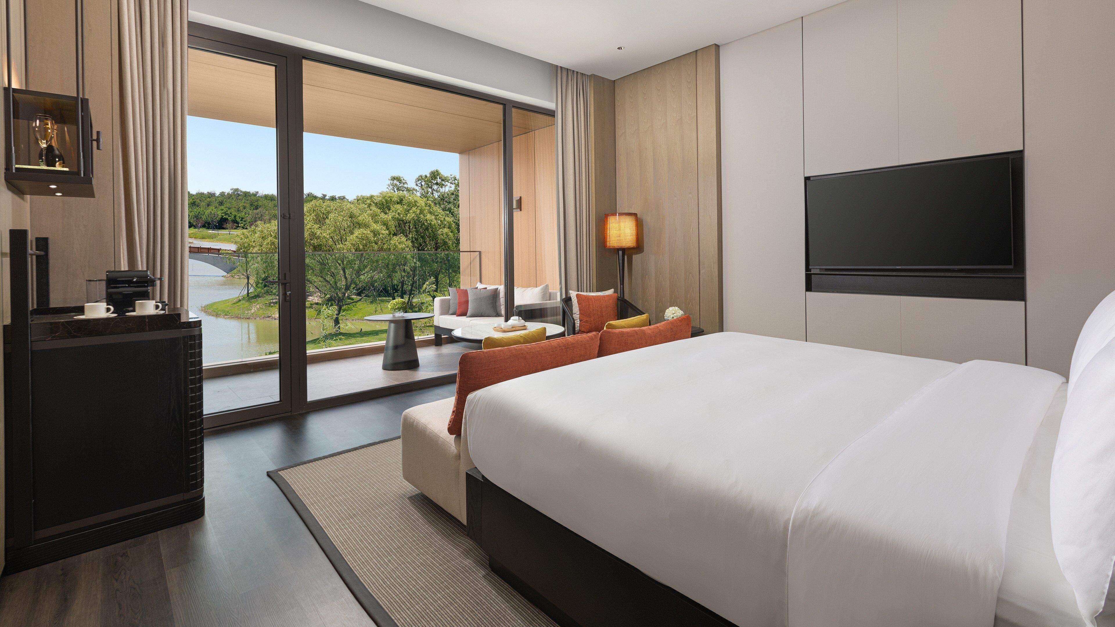 A hotel room with a large bed, a desk with a coffee maker, a TV, and a view of a garden through a sliding door.