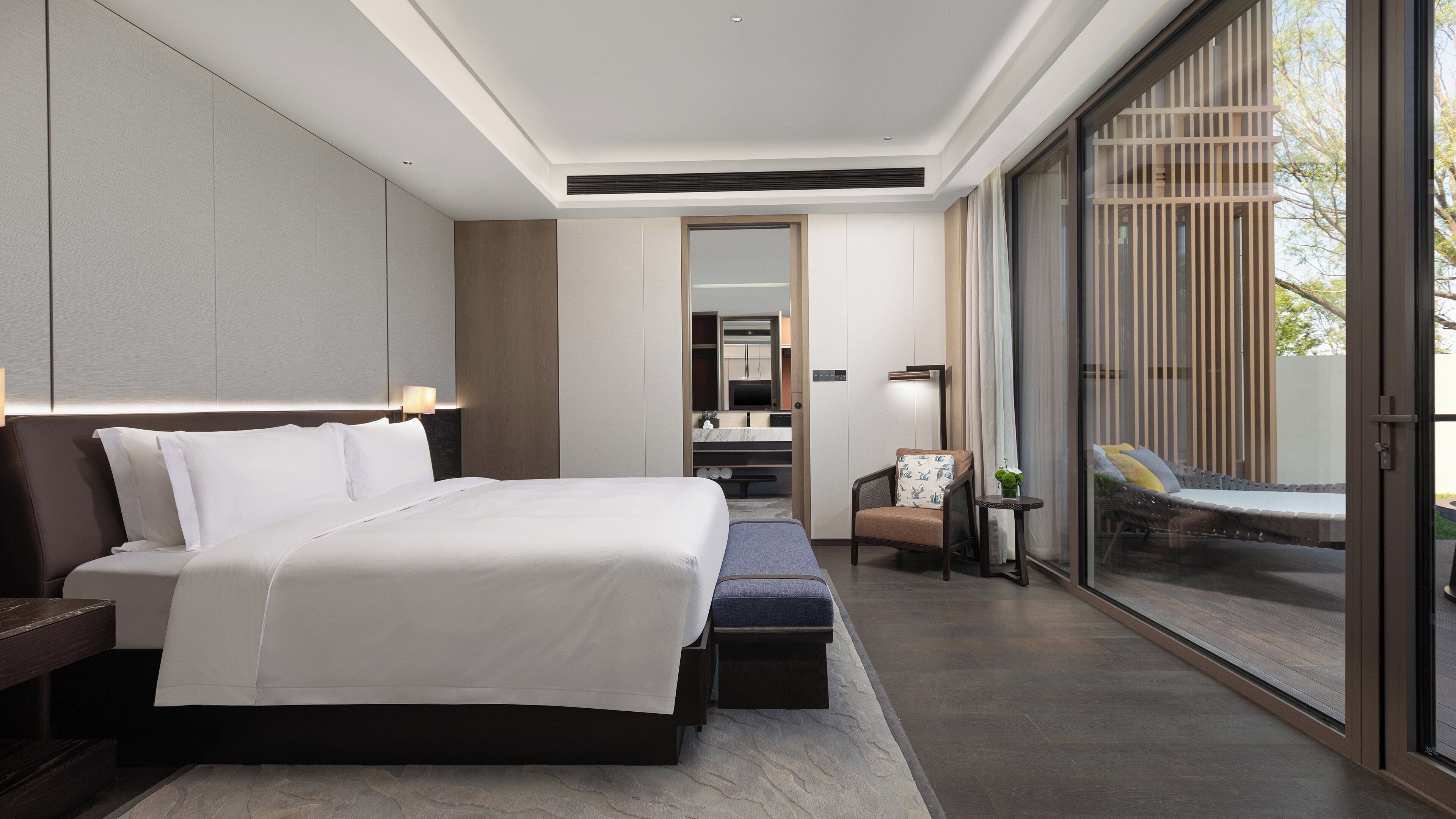 A modern hotel room with a large bed, a seating area, a small table, and a view through a glass door.