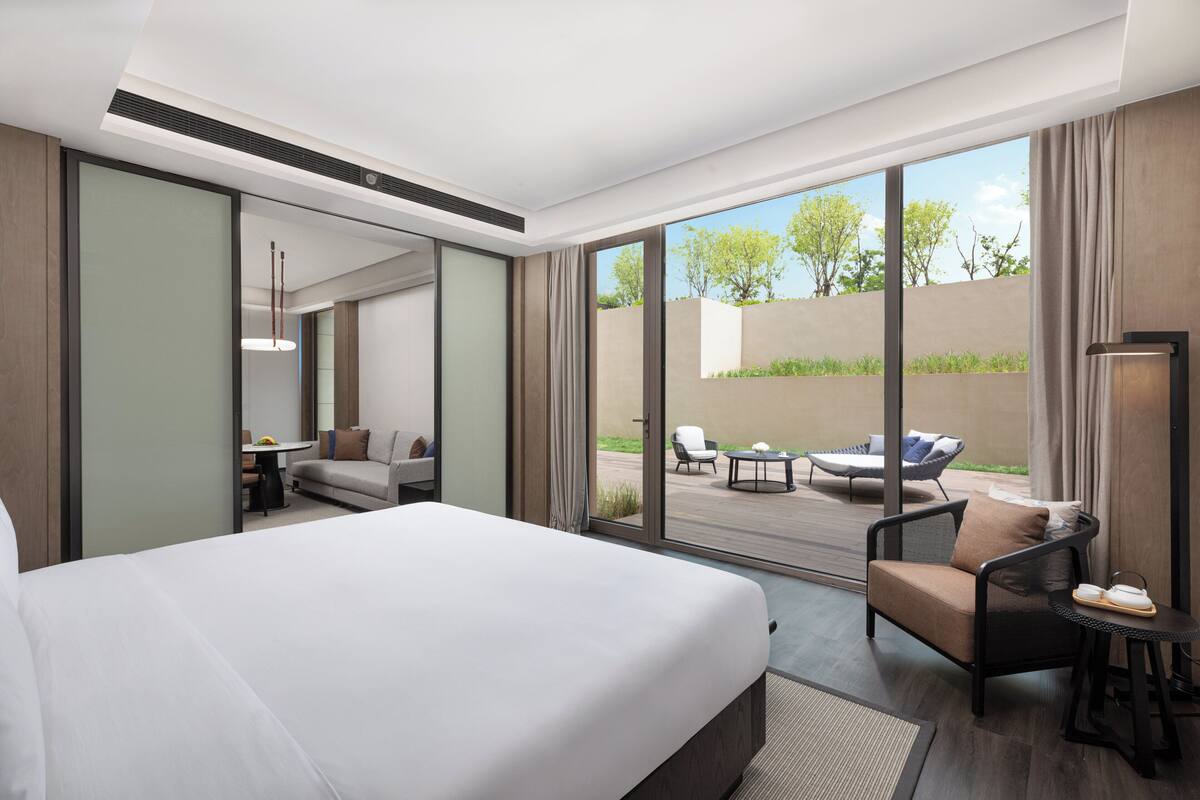 A modern hotel room with a large bed, a seating area with a sofa and armchairs, a small table, and a view of the outdoor patio.