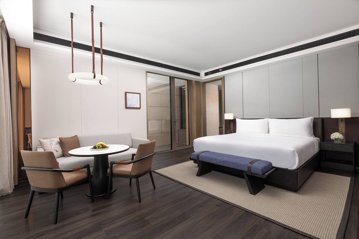 A modern hotel room with a large bed, a round dining table, two chairs, a sofa, and a small table with fruit.