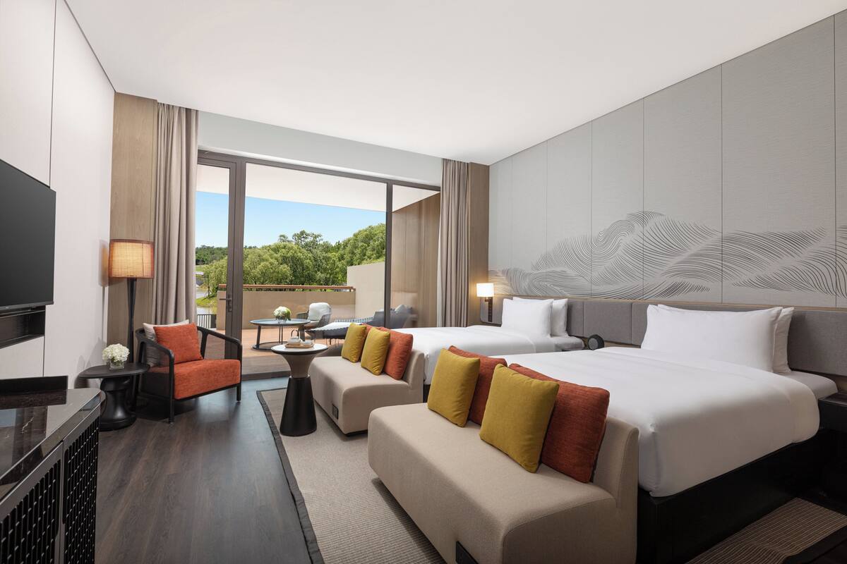 A hotel room with two beds, a sofa, a TV, and a balcony with a view of trees.