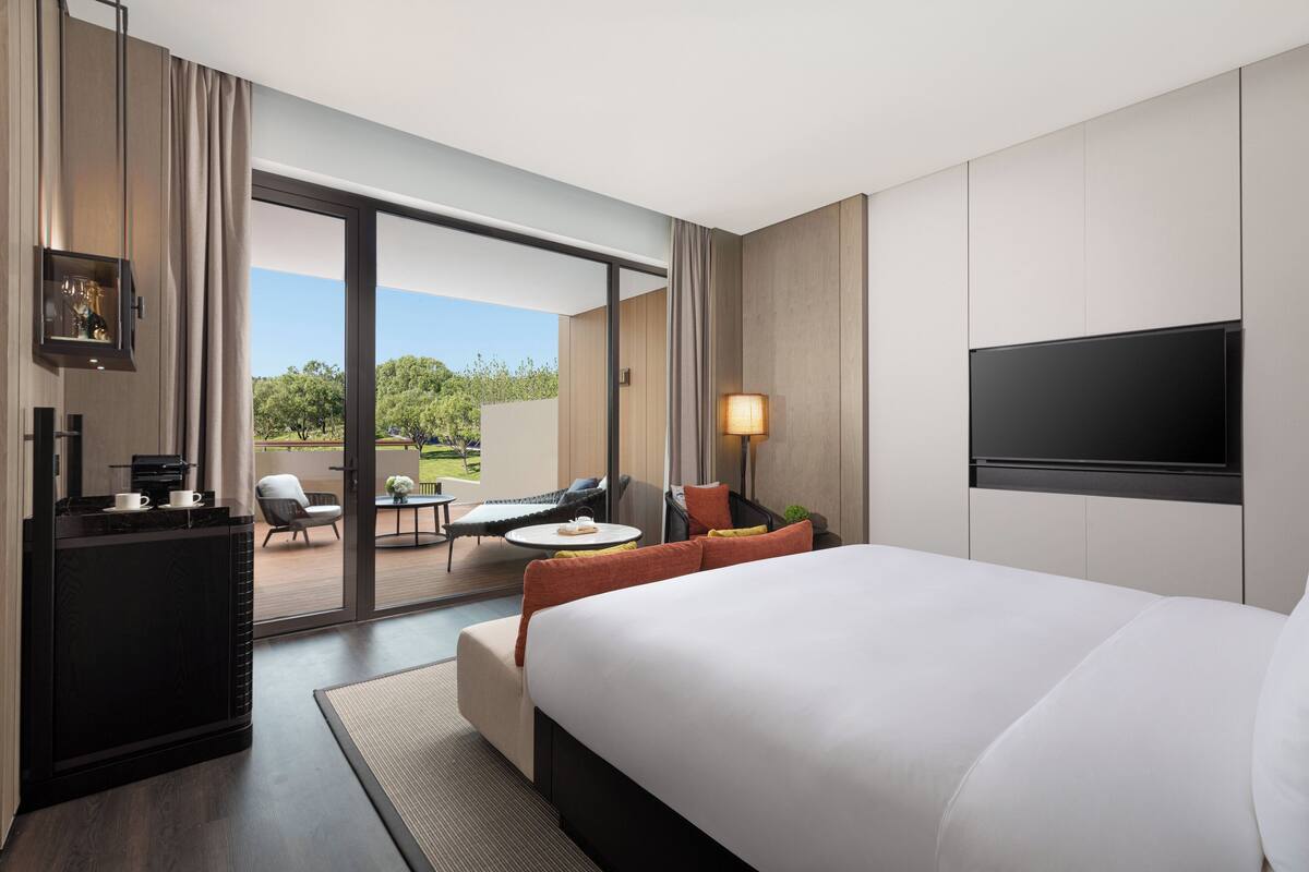 A modern hotel room with a large bed, a flat-screen TV, a desk with a coffee machine, and a balcony with outdoor seating.