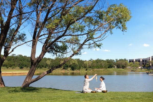 Two people sitting on grass by a lake, under a tree.