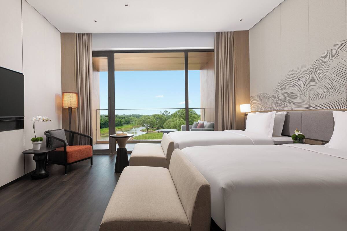 A modern hotel room with two beds, a flat-screen TV, a desk, and a view of a river and greenery.