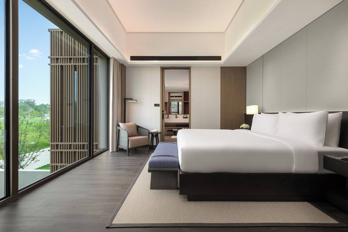 A modern hotel room with a large bed, a seating area with a chair and a small table, a large window with a view of greenery, and a bathroom visible through an open door.