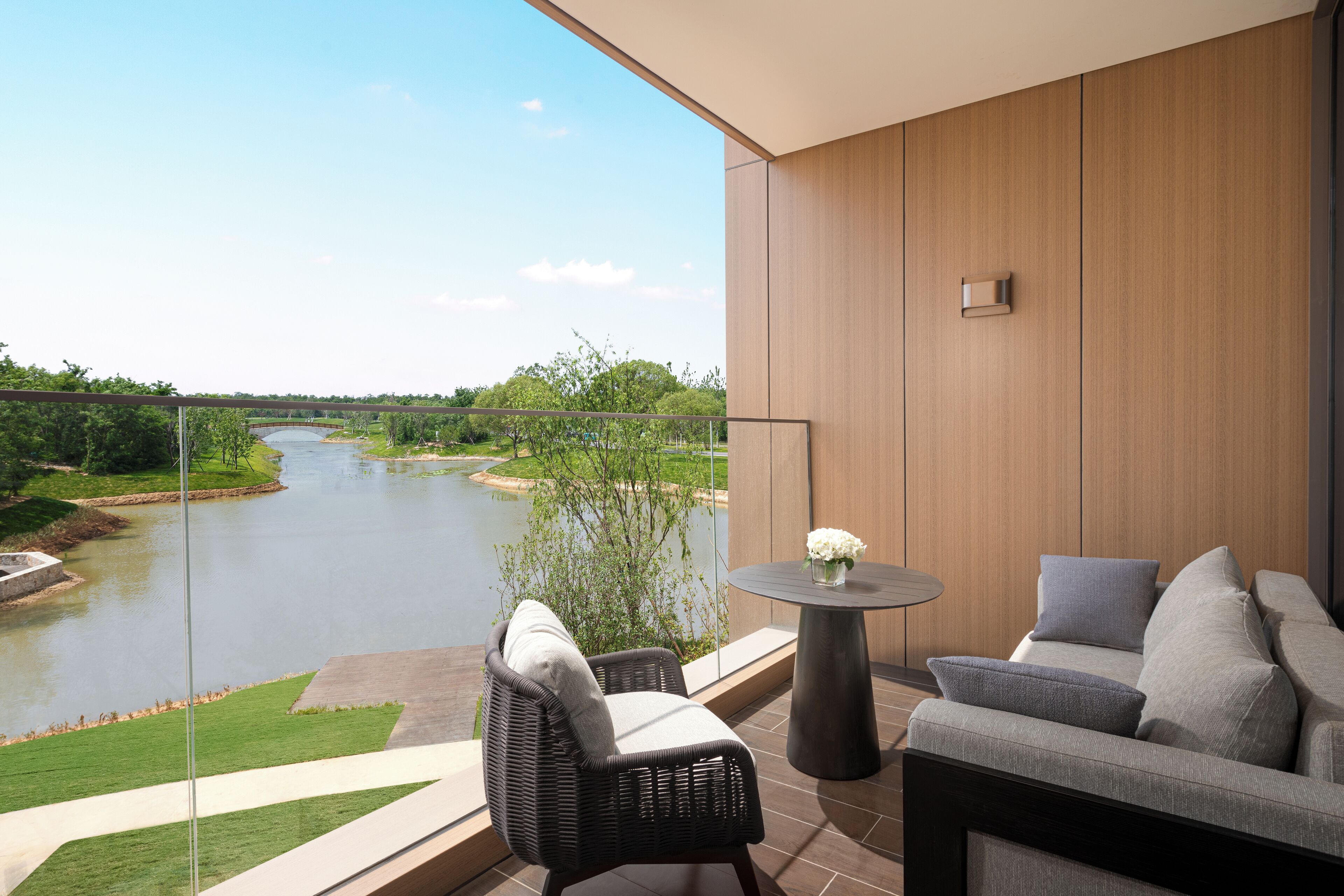 A balcony with a view of a river, a wicker chair, a grey sofa, and a small round table with a vase of flowers.