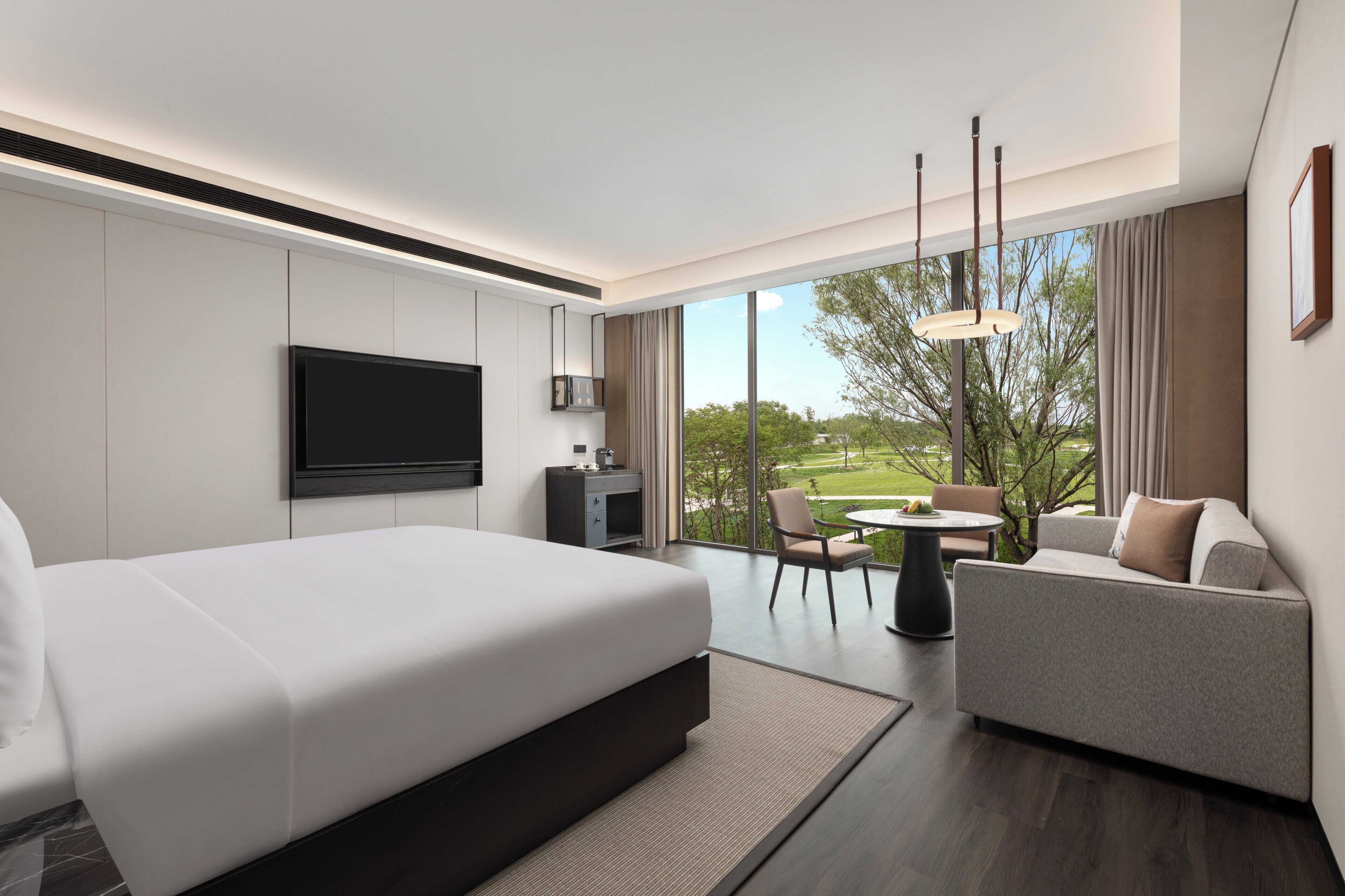 A modern hotel room with a large bed, a sofa, a dining table, and a TV mounted on the wall.