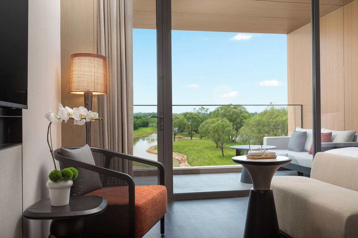 A modern hotel room with a balcony, a sofa, a chair, a table, and a TV.