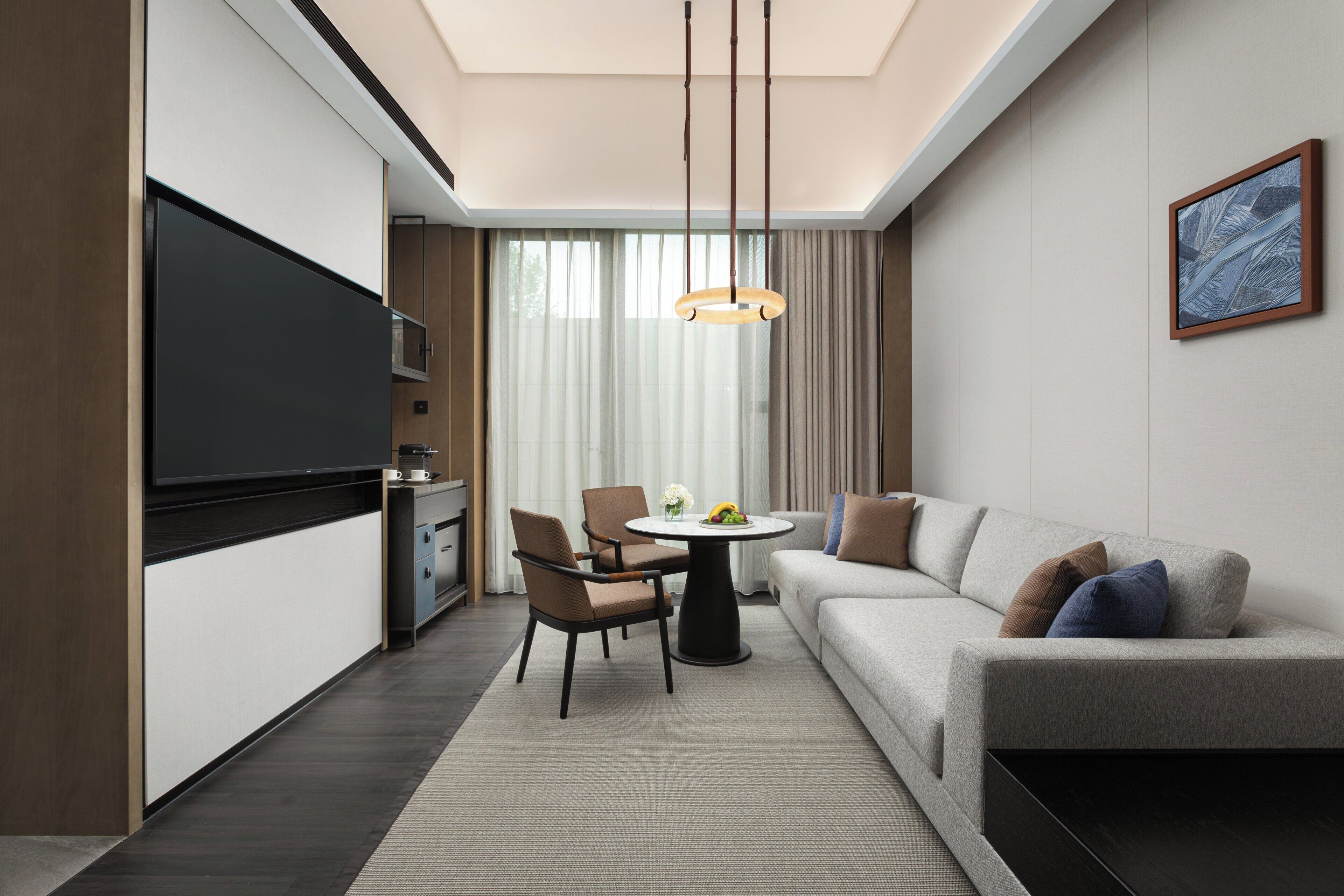 A modern hotel room with a flat-screen TV, a dining area with a table and chairs, a sofa, and a large window with curtains.