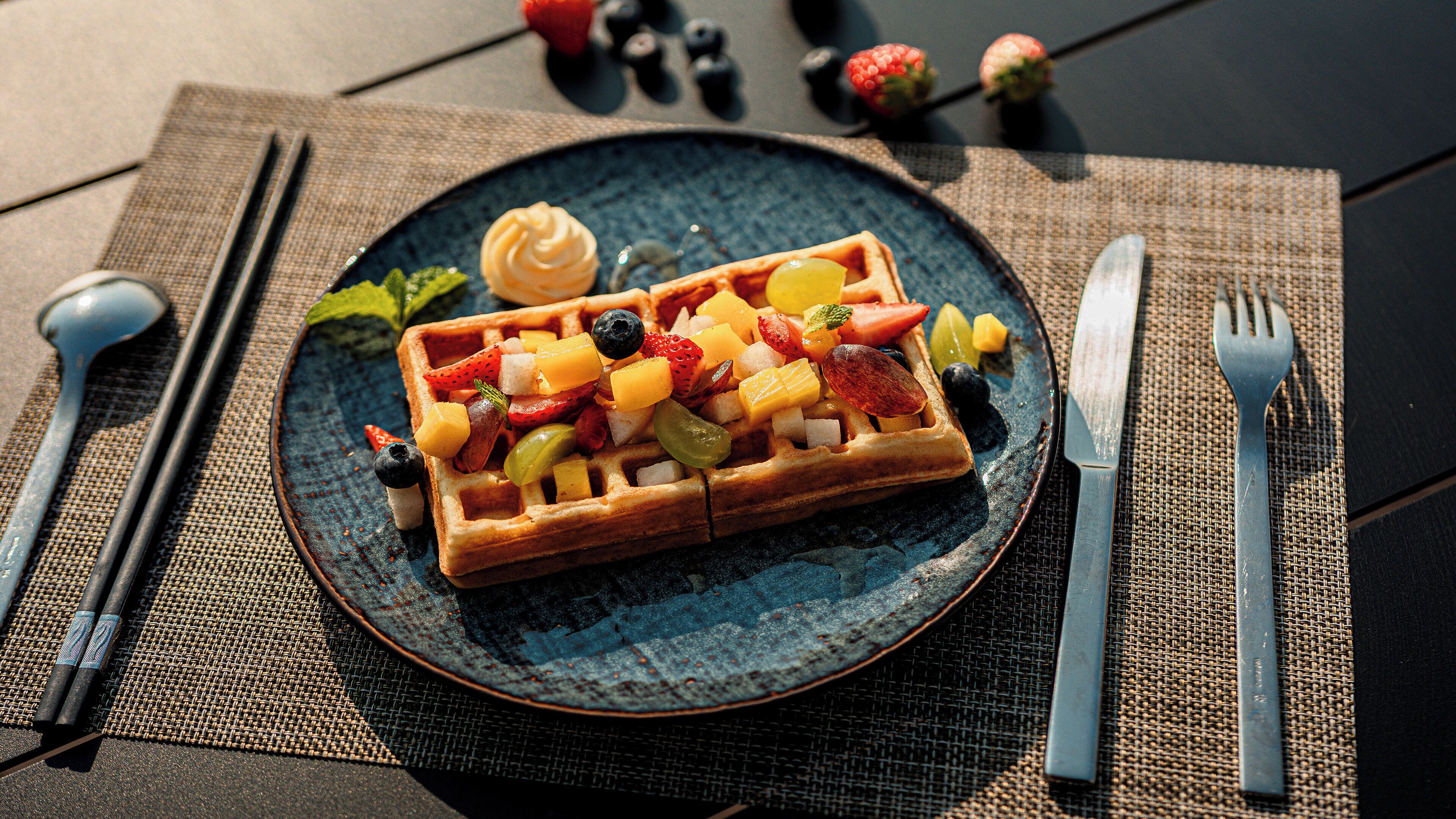 A plate with a waffle topped with fruits and a dollop of cream, accompanied by cutlery and a napkin on a woven placemat.