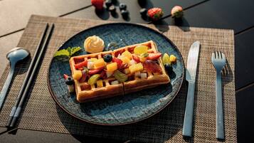 A plate with a waffle topped with fruits and a dollop of cream, accompanied by cutlery and a napkin on a woven placemat.