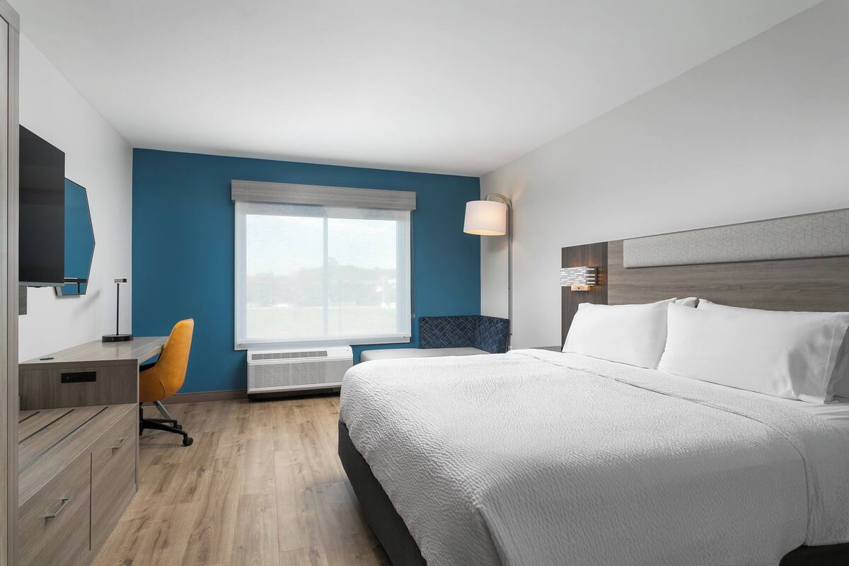 A modern hotel room with a large bed, a desk with a chair, a window with blinds, and a wall-mounted lamp.