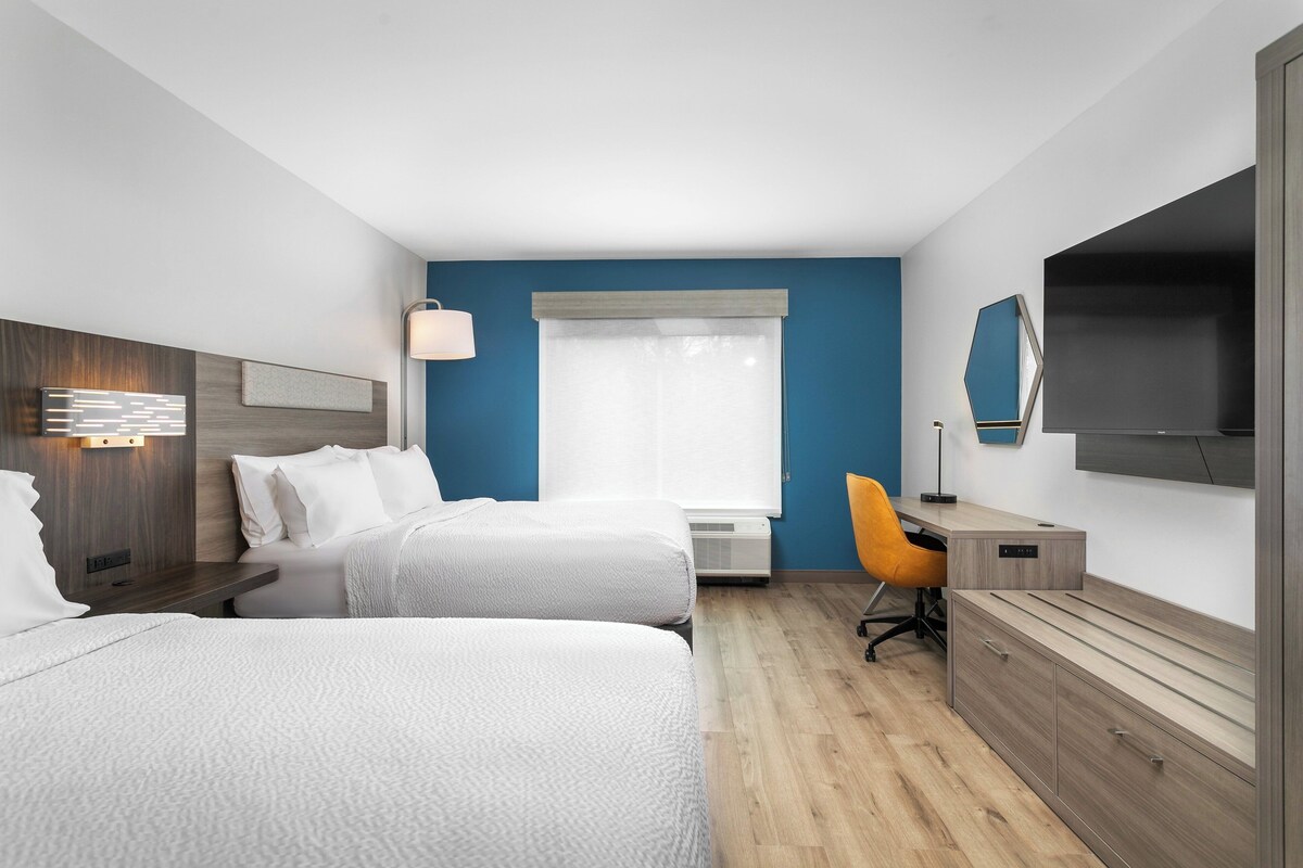 Hotelkamer met twee bedden, een bureau, een stoel, een televisie en een raam met jaloezieën.
