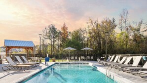 Une piscine rectangulaire entourée de chaises longues et de parasols, elle-même bordée d’arbres et d’un belvédère.