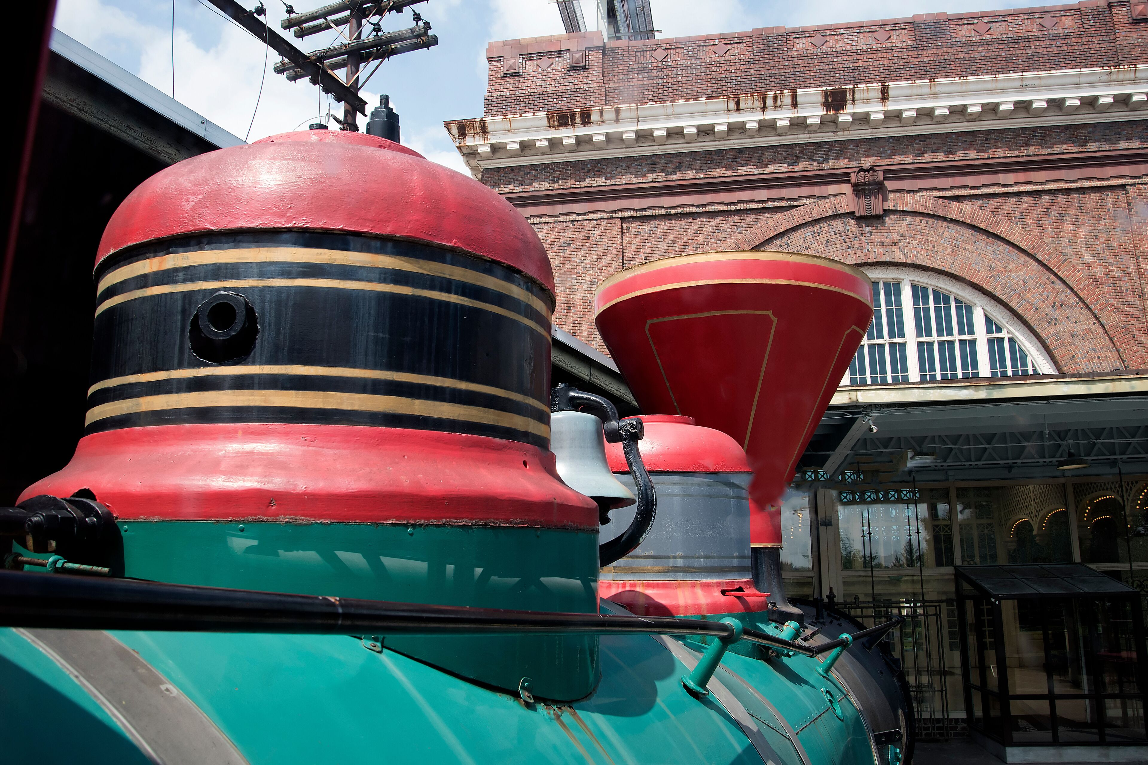 Une locomotive à vapeur, avec une partie avant rouge et noire, une carrosserie verte et une cheminée rouge, est garée devant un bâtiment en briques doté de grandes fenêtres et d’une structure en verre.