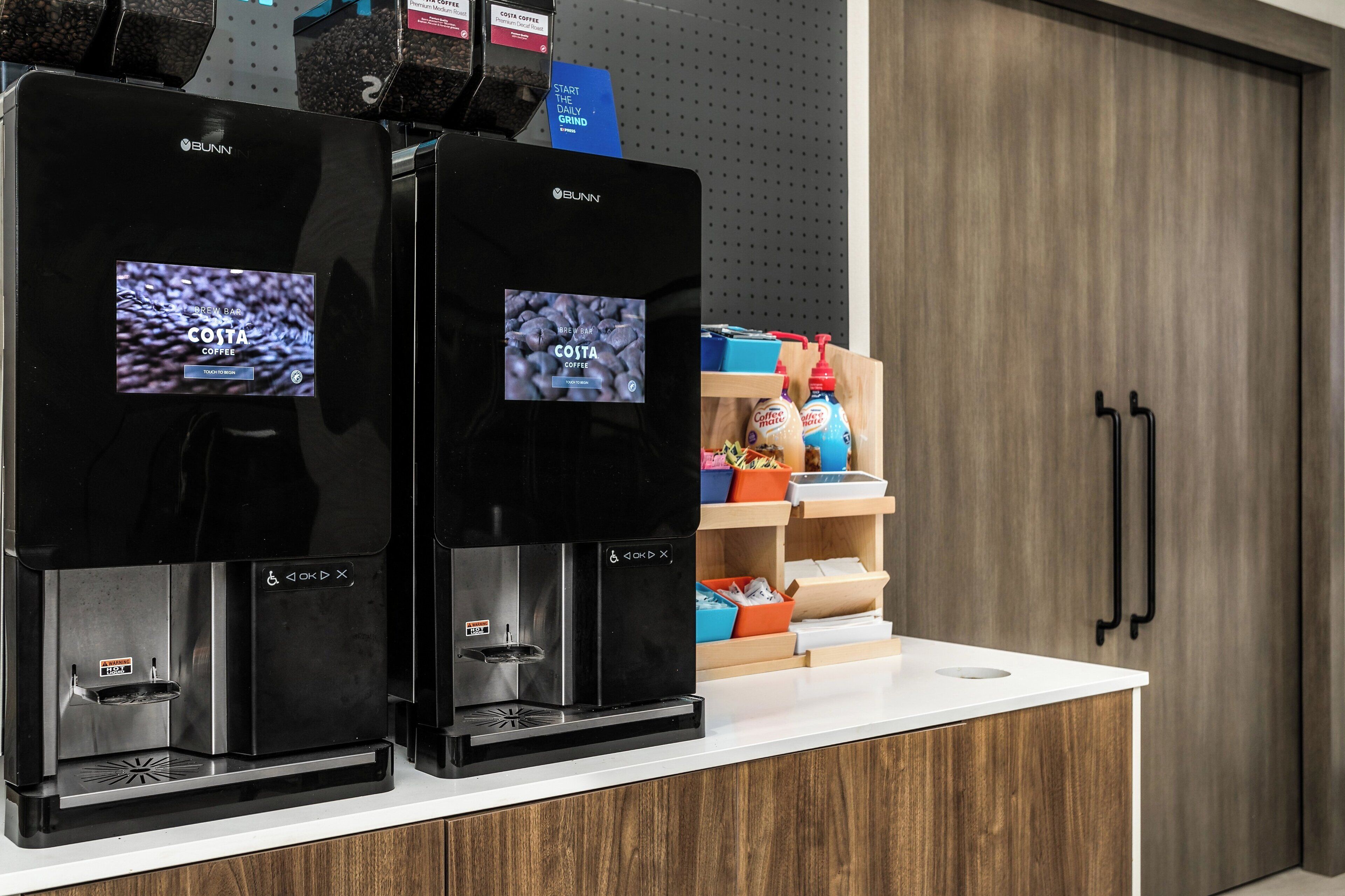 Two Bunn coffee machines on a counter with a wooden cabinet and shelves in the background.