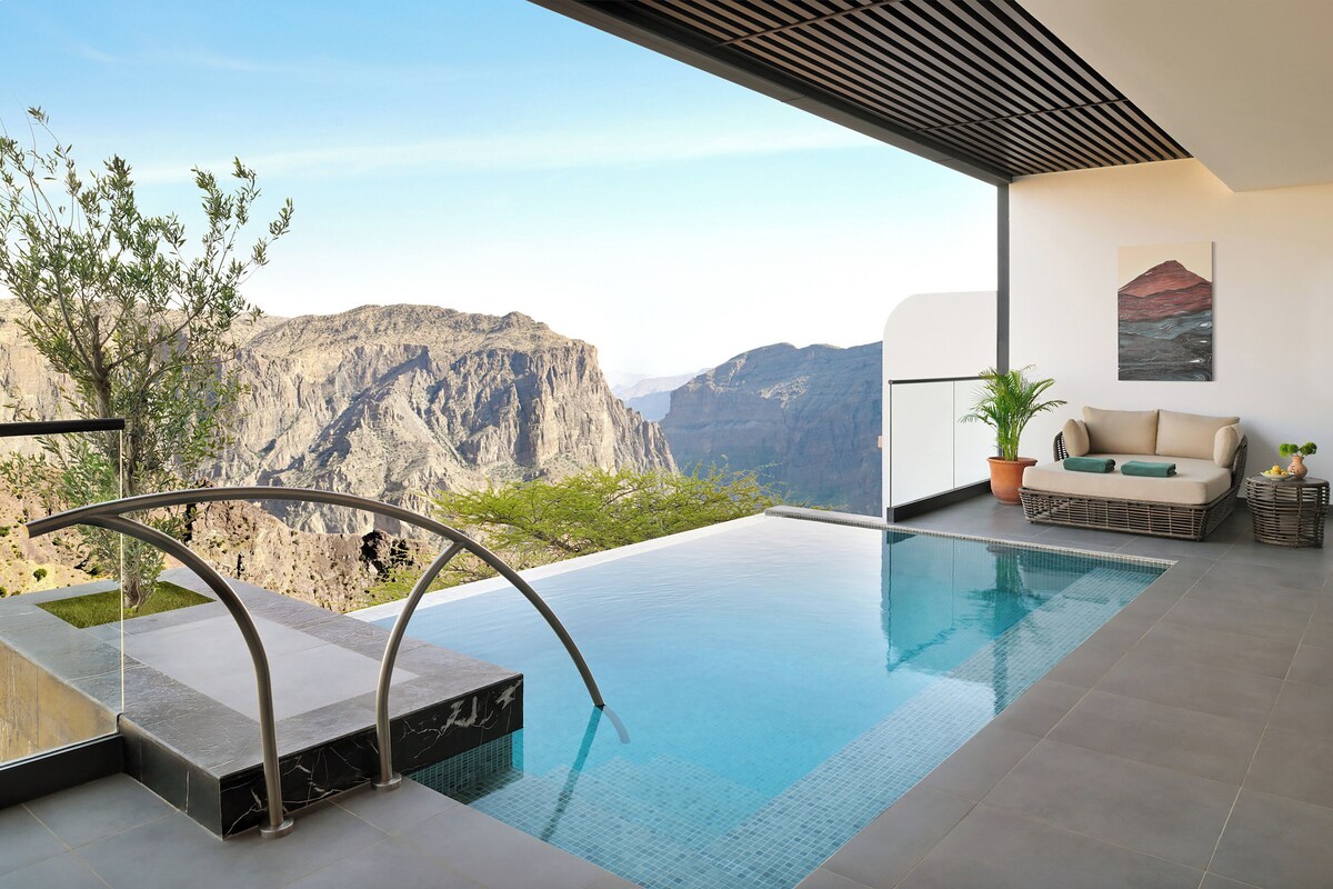 Une piscine à débordement moderne offrant une vue sur les montagnes, un espace de détente avec un canapé et des plantes en pot, et un design architectural minimaliste.