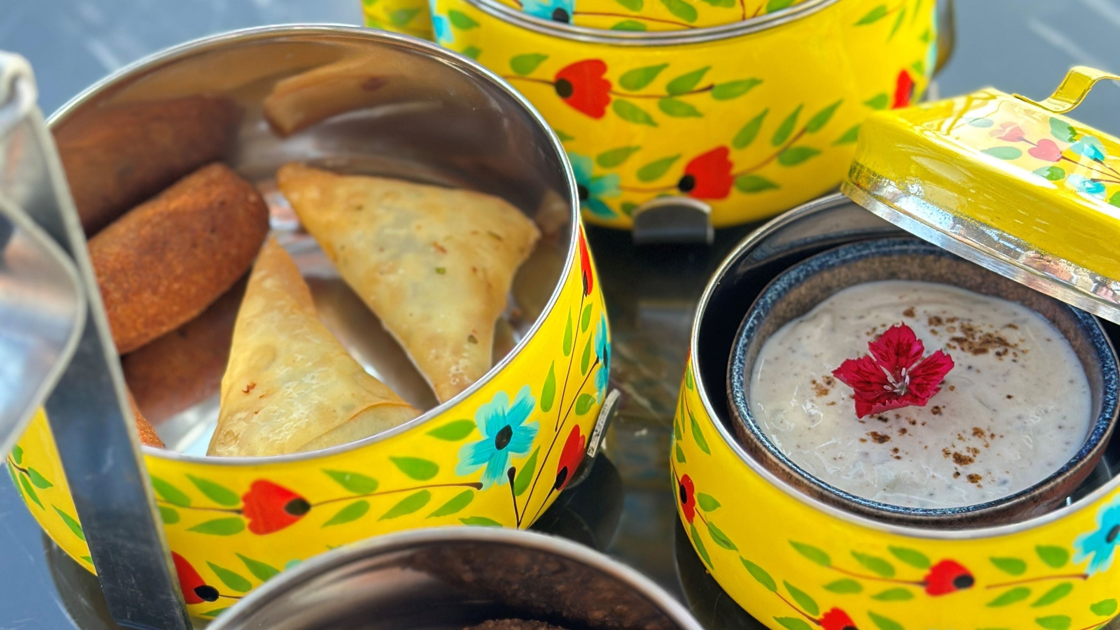 A close-up of a meal with samosas and a bowl of white soup garnished with a red flower, served in colorful, floral-patterned containers.