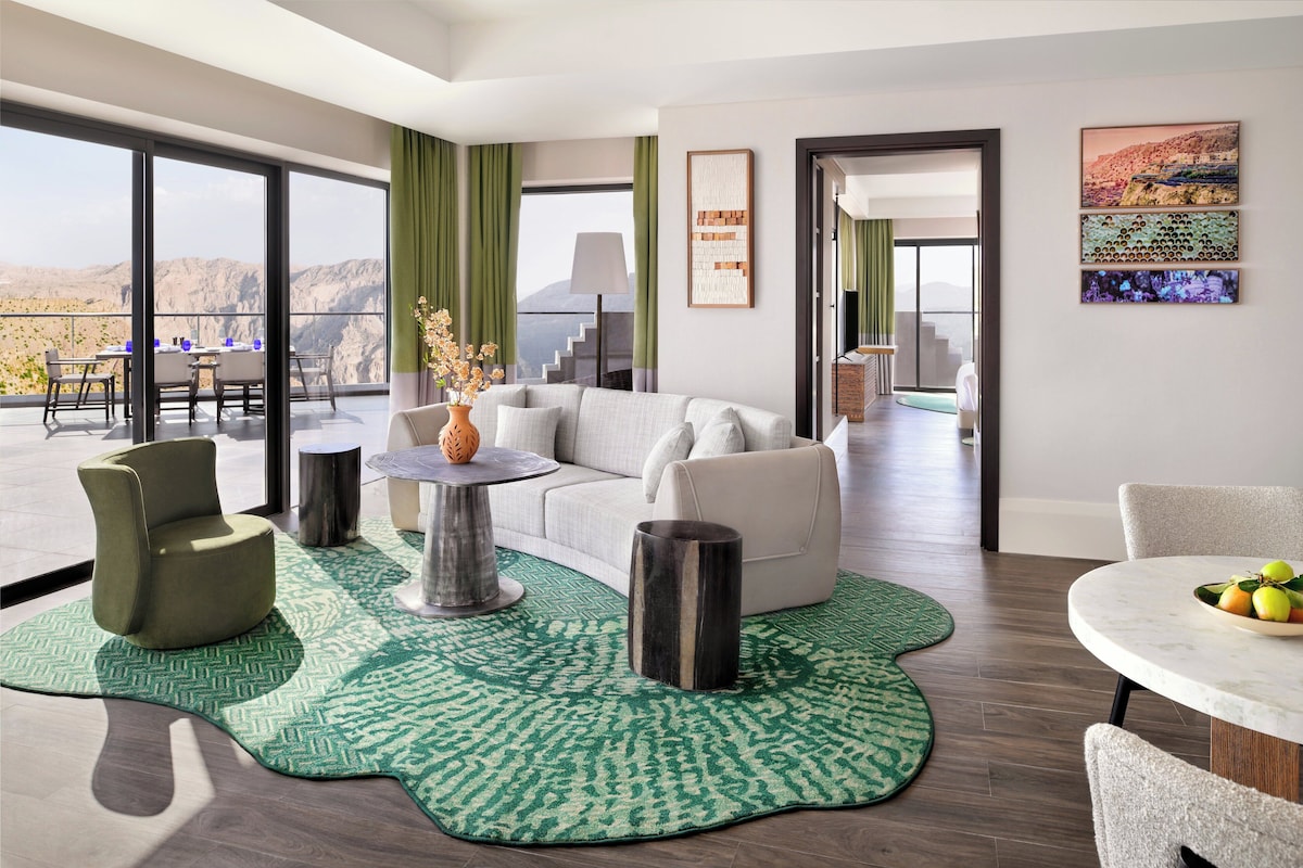 A modern living room with a sofa, chairs, a coffee table, and a dining table with fruit. There is a large window with a view of mountains.
